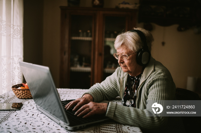 Side view of a senior caucasian woman with headphones while sitting at a table in front of a laptop during quarantine COVID - 19 coronavirus