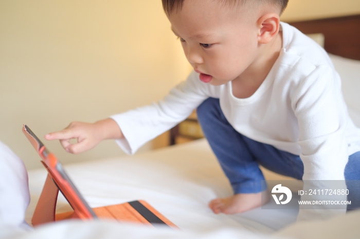 Cute little Asian 2-3 years old toddler boy child sitting in bed watching a video from tablet pc. Kids playing with tablet computer, Gadget-addicted children, pseudo-autism concept