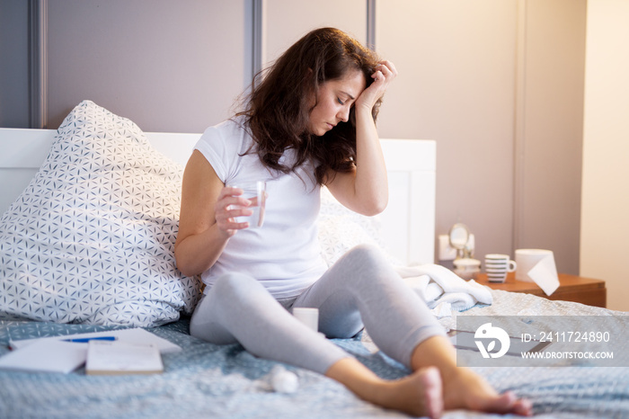 Tired ill middle aged woman holding hand on the forehead while having the glass of water in another hand and paper wipes are around her on the bed.