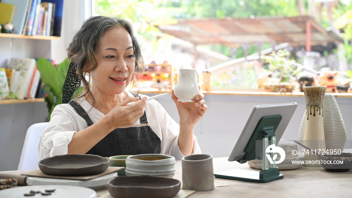 Concentrated middle aged woman painting pottery bowl in workshop. Indoors lifestyle activity, handicraft, hobbies concept