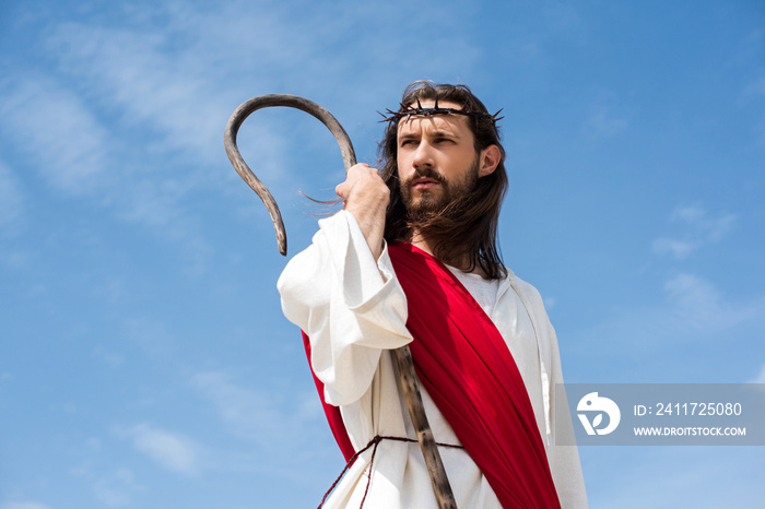 Jesus in robe, red sash and crown of thorns standing with wooden staff against blue sky and looking away