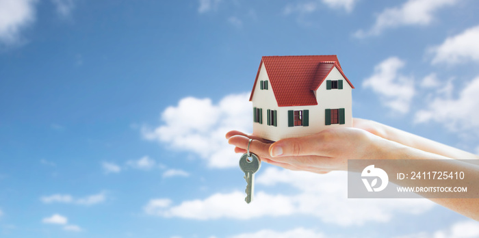 mortgage, real estate and property concept - close up of hands holding house model and home keys over blue sky and clouds background