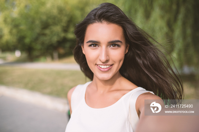 Self portrait of beautiful young brunette smiling woman on nature and sky background in the park. Travel. Selfie. Instagram