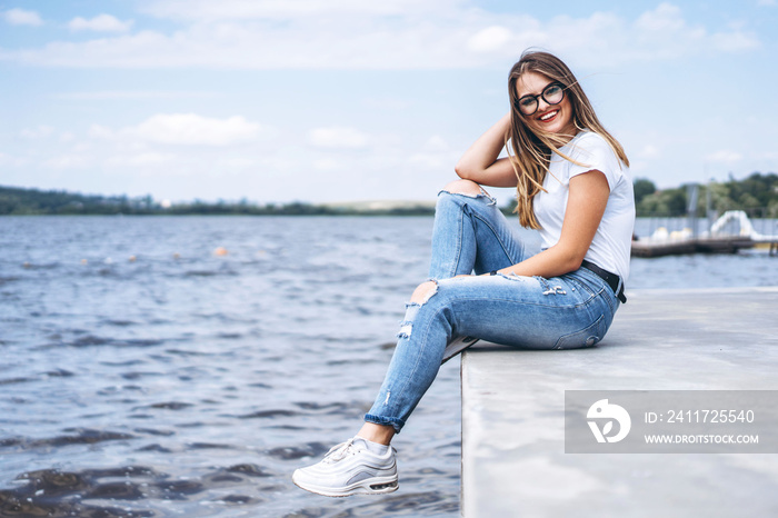 Young woman with long hair in stylish glasses posing on the concrete shore near the lake. Girl dressed in jeans and t-shirt smiling and looking at the camera