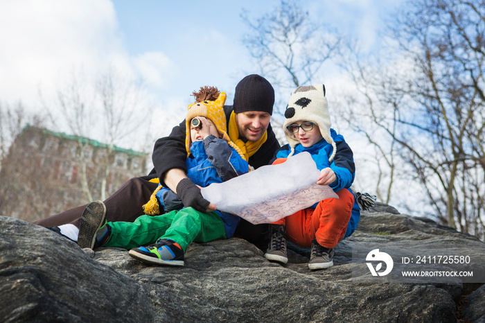 Father with boys reading map while sitting on rock against sky