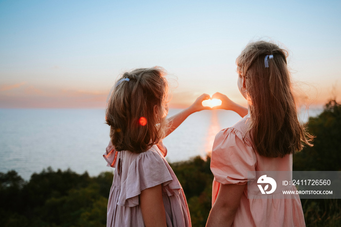 Two teen sisters with loose hair wearing pastel dresses from behind looking at beautiful summer sunset over the sea and making heart gesture with their hands. Sisterhood