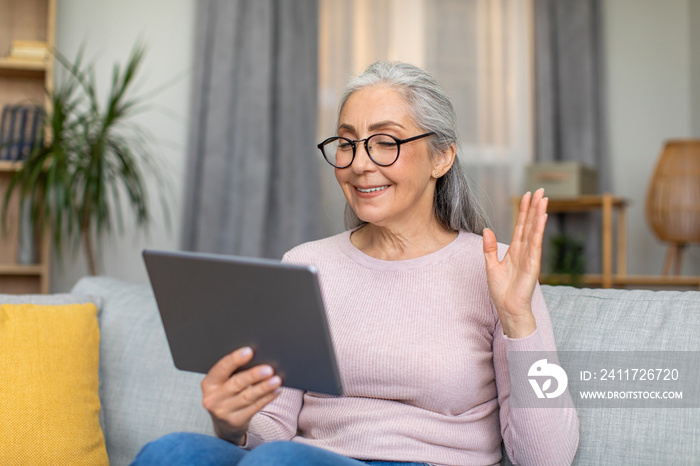 Glad european mature woman with gray hair in glasses waves her hand and looks at tablet, have meeting