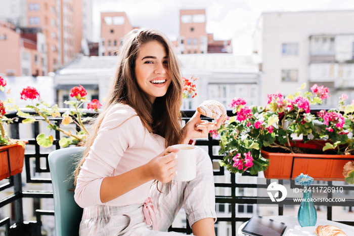 Portrait pretty girl  having breakfast on balcony surround flowers in the sunny morning in city. She holds a cup, croissant, smiling to camera