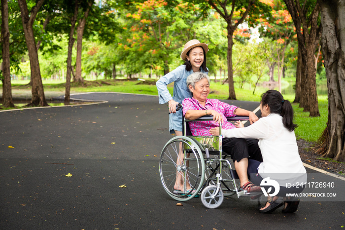 Asian senior woman in wheelchair with little child girl support,enjoy on walking,grandmother with her daughter,granddaughter having fun talk,hug,laughing together in park,elderly care,family concept