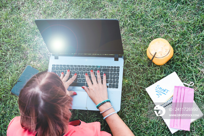 Woman 40 years old working outdoors with laptop while making healthy breakfast - Female entrepreneur or influencer lying down on lawn typing on pc during the end of coronavirus outbreak - Top view