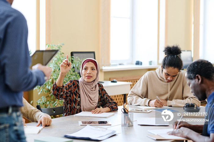 Muslim woman attending language lessons for immigrants raising hand to ask teacher question about task