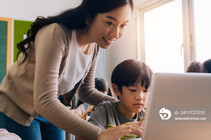 Young adult smiling beautiful Asian teacher helping elementary student boy with laptop in computer classroom. Information Technology class in primary school concept.