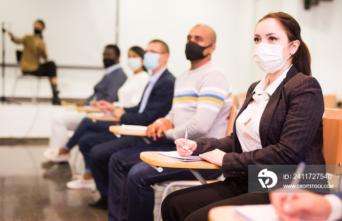 Young focused woman in protective face mask sitting and listening to speaker at business conference