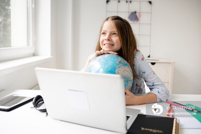 beautiful, young, blond girl is sitting in front of laptop holding globe in her hands and dreaming about vacation and end of pandemic and covid 19