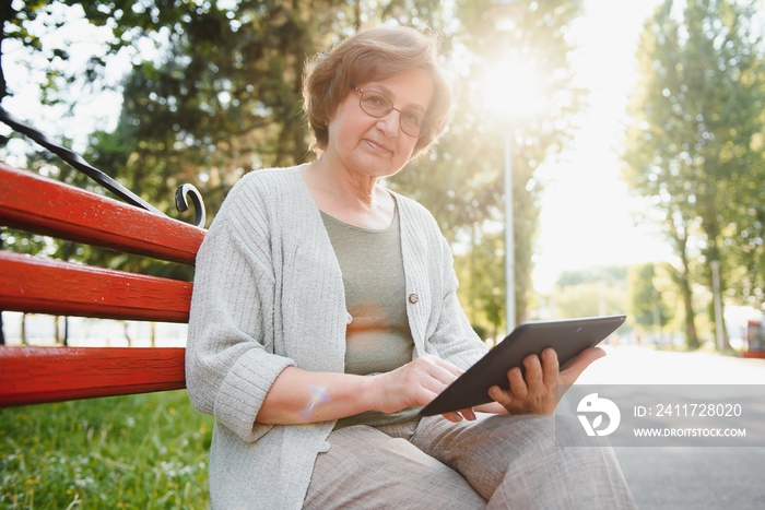 Positive confident old lady posing with tablet in park. Senior grey haired woman in casual sitting on park bench and using tablet. Wireless connection concept