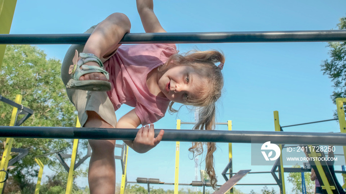 Little girl climbs gymnastic ladder on open sports ground on outside. Cute little girl crawls on vertical sports ladder in city park on sun day.