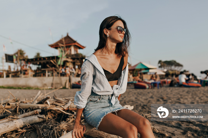 Happy attractive lovely woman with dark short hairstyle wearing denim skirt and light shirt sitting on the ocean shore and enjoying ocean view on sunset