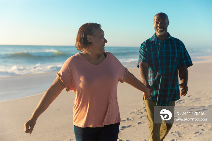 Smiling african american senior woman holding hand of man walking at beach