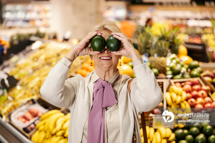 A goofy old woman is playing with avocado and making faces at the supermarket.