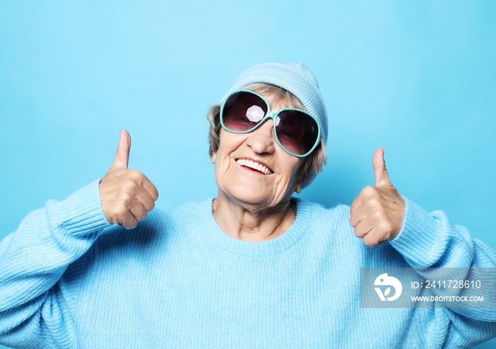 Lifestyle, emotion and people concept: Funny old lady wearing blue sweater, hat and sunglasses showing victory sign.