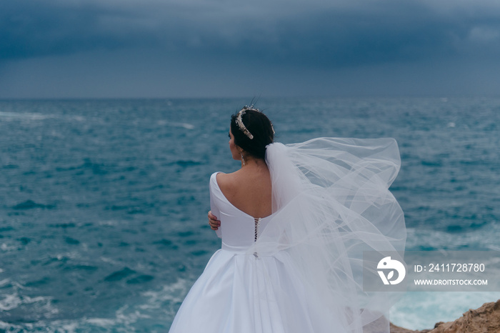 Back view of brunette bride in white wedding dress and bridal veil on a cloudy day. Romantic beautiful bride posing near the sea with waves