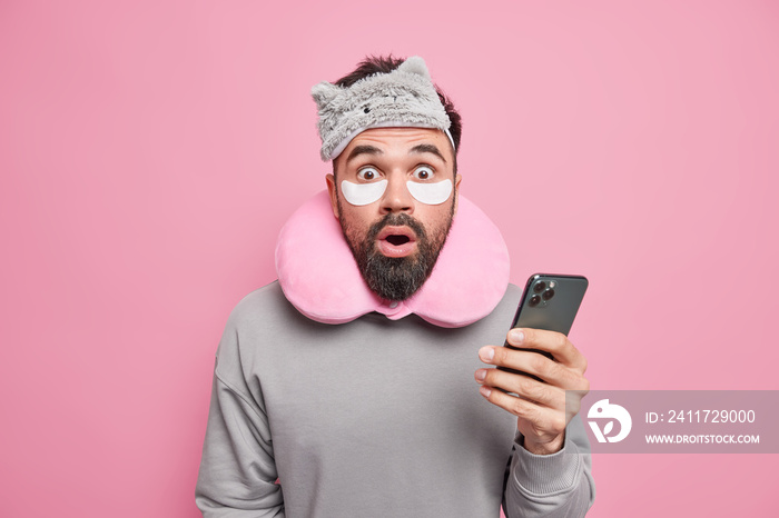 Amazed bearded man reacts on astonishing news looks with disbelief stands speechless holds breath uses smartphone surfs internet prepares for having nap wears sleepmask neckpillow isolated on pink