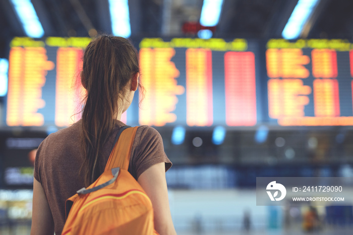 Young woman with small backpack as a hand luggage in international airport looking at the flight information board, checking her flight