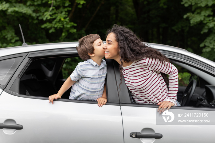 Happy little boy kissing millennial mom in open car window, outdoor in summer, free space