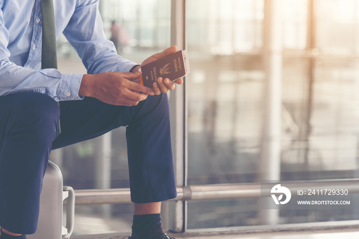 Young traveling businessman, sitting on baggage handing passport - airport security concept. Man holding a Thai passport waiting for his flight in airport terminal hall.