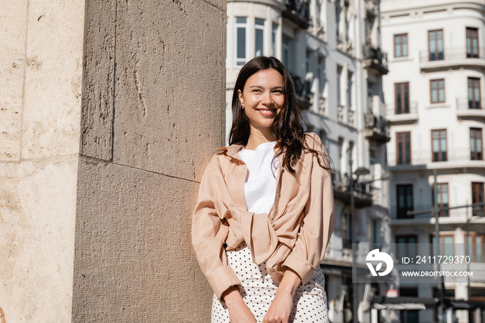 happy woman in beige shirt standing on city street near wall.