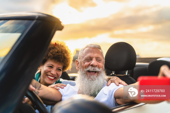 Happy senior couple having fun on new convertible car roadtrip taking selfie with mobile smartphone - Travel people lifestyle concept