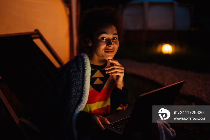 Young african woman working on laptop while sitting on chair near glamping tent in nature