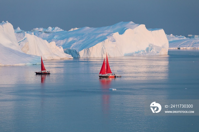 Little red sailboat cruising among floating icebergs in Disko Bay glacier during midnight sun season of polar summer. Ilulissat, Greenland.  Expedition ship with iceberg in Antarctic sea