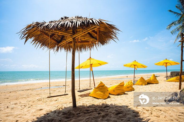 Yellow umbrella with yellow beach chairs on the beach with brown sand, blue sea water, clear blue sky and coconut trees in background.