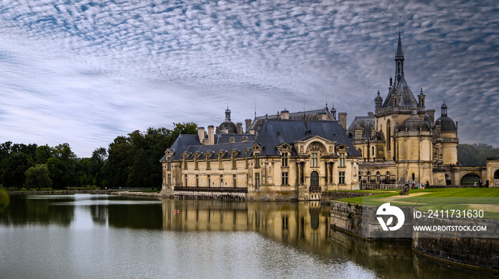 Panoramic view of the Château de Chantilly reflected in the water of the river with light clouds in the sky.