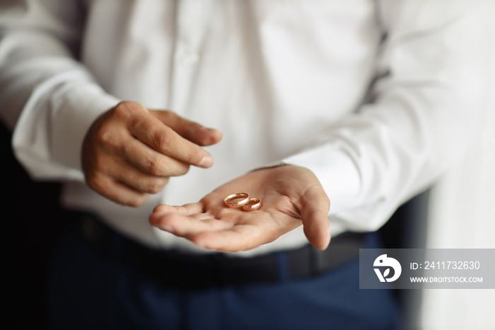 The man the groom in a white shirt and blue trousers holds on his palm wedding gold rings by the window. Groom morning, close-up without a face only hands