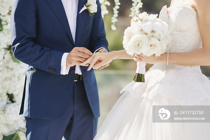 Hands with wedding rings. Modish groom putting a golden ring on the bride’s finger during the wedding ceremony. Loving couple, a girl in a wedding dress and handsome man in a stylish blue suit