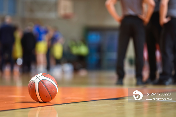 Basketball ball on the parquet with team and referee in the background.