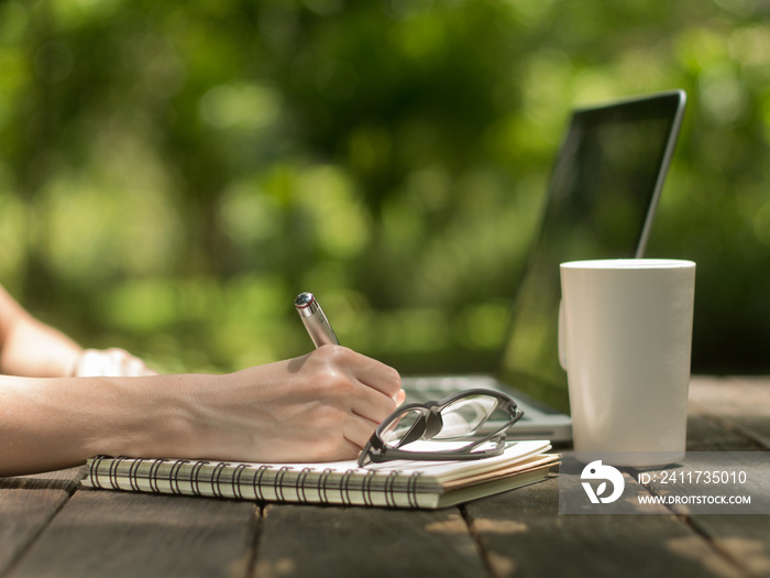 Hand of woman writing on a book with white coffee cup on the old wooden desk in the garden.