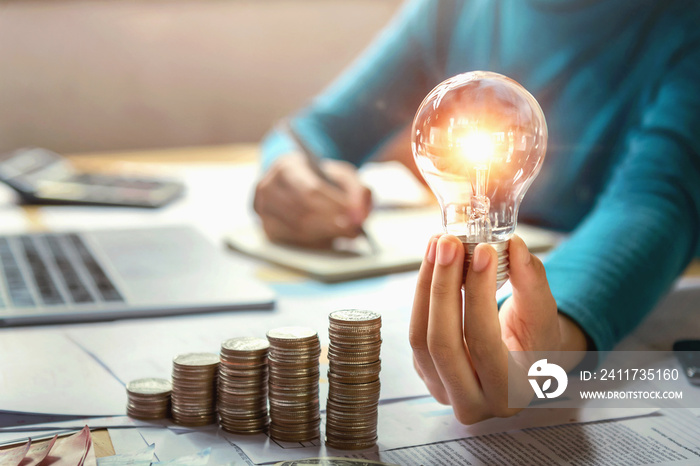 business woman hand holding lightbulb with coins stack on desk. concept saving energy and money