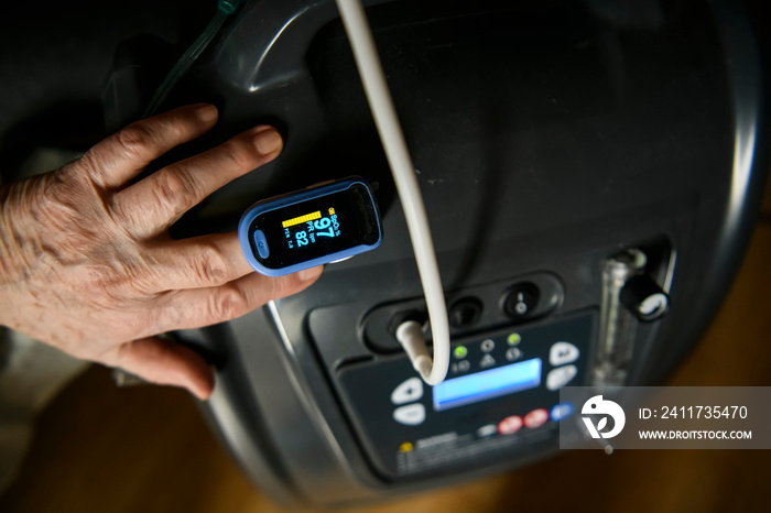 View of a hand of a woman with an pulse oximeter clipped on the index finger. Hand lies on a medical oxygen concentrator