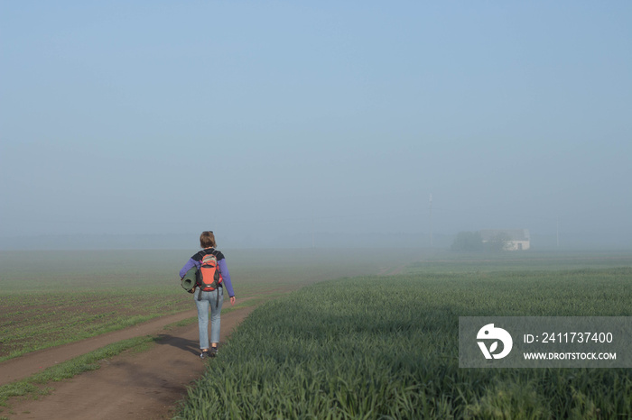 Woman traveler with backpack walks in beautifyl atmospheric misty morning  a house among fields in the background