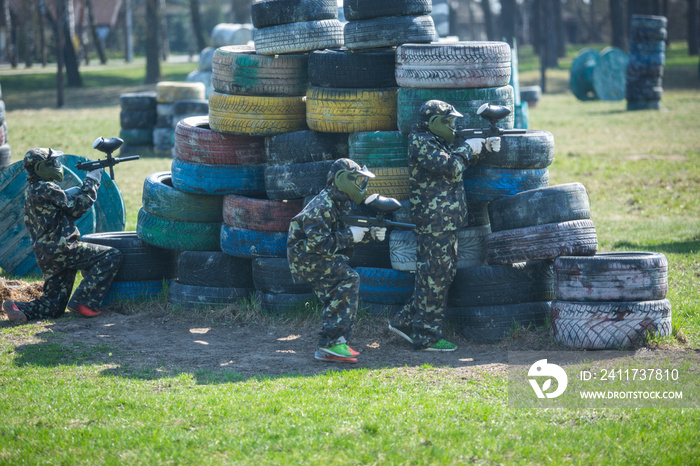 Paintball players in camouflage uniform and protective mask with gun on the field, shoot into enemies in the summer