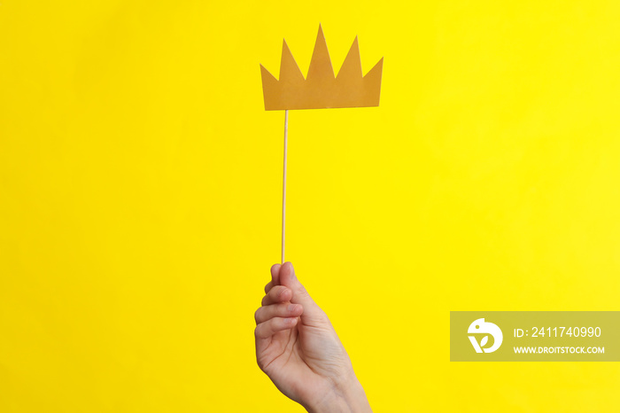 Hand holds paper cut golden crown with stick on yellow background. Party concept