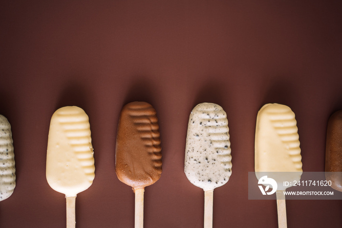 Line of milk and white chocolate popsicles on brown background, top view.