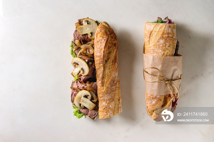 Opened and tied beef baguette sandwich with champignon mushrooms, green salad, fried onion over white marble background. Flat lay, space