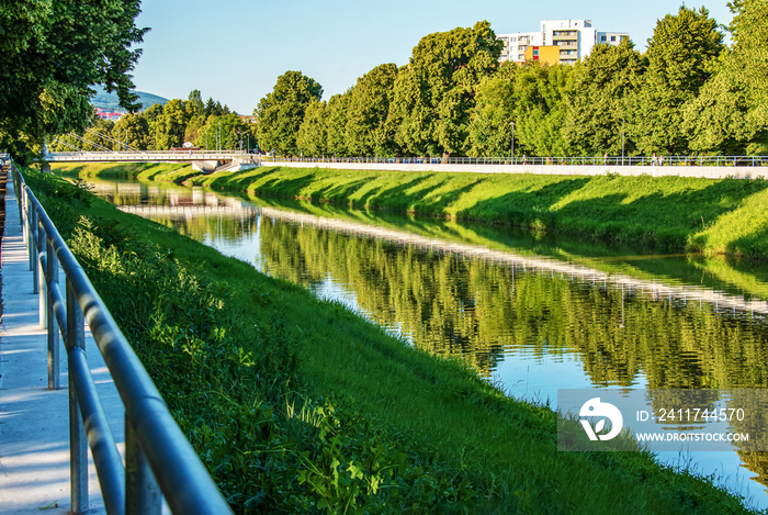 The river flows through the city. On the calm surface of the river banks are reflected. Slovak river Nitra on a bright sunny day.