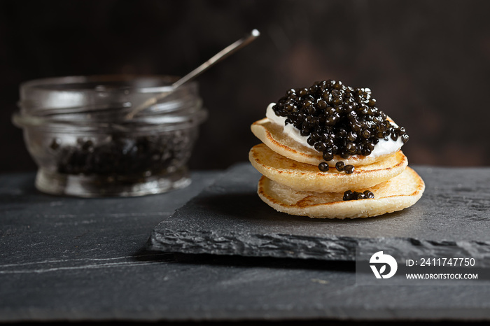 Black sturgeon caviar on small pancakes blinis with sour cream and a glass jar with caviar on a slate