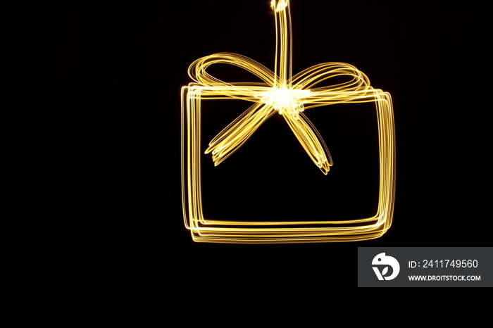 Long exposure photograph of neon gold colour in the shape of a present, gift box outline against a black background. Light painting photography.