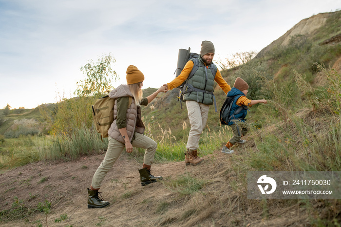 Young active family of hikers with backpacks moving upwards on top of mountain